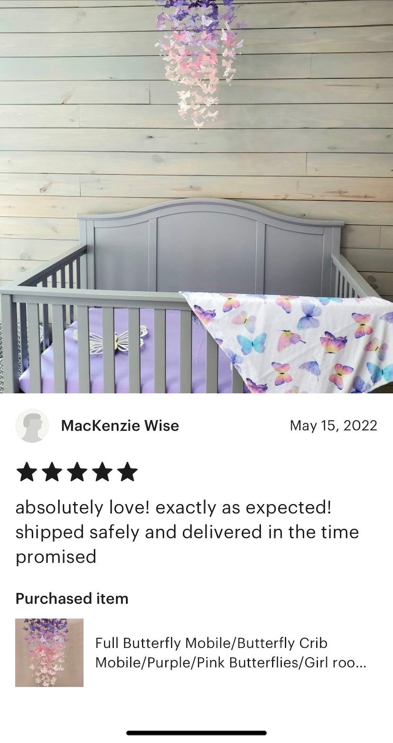 K&ouml;nnte beinhalten: Ein lila und pinkfarbener Schmetterlingsmobile h&auml;ngt &uuml;ber einem Kinderbett mit einem wei&szlig;en und lilafarbenen Schmetterlingsmuster. Das Mobile besteht aus Papierschmetterlingen und ist eine tolle Erg&auml;nzung f&uuml;r ein Kinderzimmer.