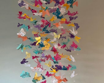 May include: A colorful paper butterfly mobile with a white circular frame. The butterflies are in various shades of pink, purple, blue, green, yellow, and white. The mobile is hanging from the ceiling.