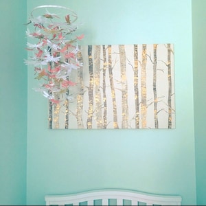 May include: A white and gold birch tree canvas print hangs on a light blue wall with a white and pink paper dragonfly mobile hanging above it.