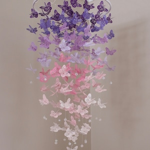 K&ouml;nnte beinhalten: Ein Papier-Schmetterlings-Mobile mit einem Farbverlauf von lila, rosa und wei&szlig;en Schmetterlingen, die an einem wei&szlig;en Metallring h&auml;ngen. Die Schmetterlinge sind in einer kaskadierenden Anordnung angeordnet, wobei die gr&ouml;&szlig;ten Schmetterlinge oben und die kleinsten unten sind. Das Mobile ist mit klaren Kristallperlen verziert.