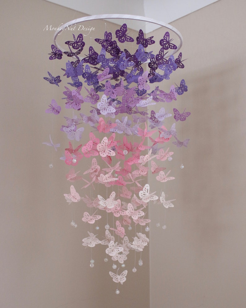 K&ouml;nnte beinhalten: Ein Papier-Schmetterlings-Mobile mit einem Farbverlauf von lila, rosa und wei&szlig;en Schmetterlingen, die an einem wei&szlig;en Metallring h&auml;ngen. Die Schmetterlinge sind in einer kaskadierenden Anordnung angeordnet, wobei die gr&ouml;&szlig;ten Schmetterlinge oben und die kleinsten unten sind. Das Mobile ist mit klaren Kristallperlen verziert.