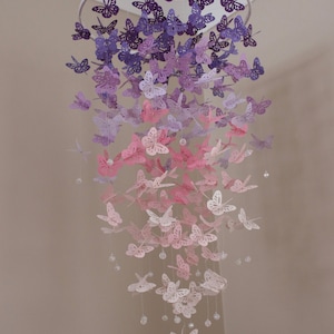 K&ouml;nnte beinhalten: Ein Papier-Schmetterlings-Mobile mit einem Farbverlauf von lila, rosa und wei&szlig;en Schmetterlingen, die an einem wei&szlig;en Metallring h&auml;ngen. Das Mobile h&auml;ngt an einer Decke.