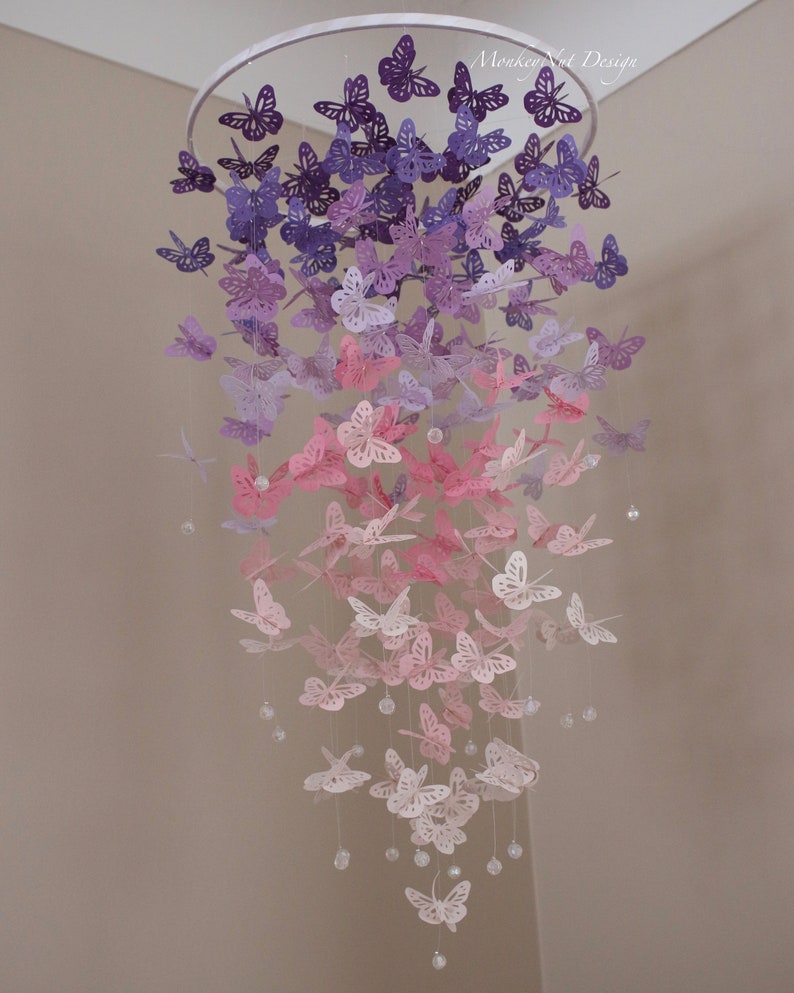 K&ouml;nnte beinhalten: Ein Papier-Schmetterlings-Mobile mit einem Farbverlauf von lila, rosa und wei&szlig;en Schmetterlingen, die an einem wei&szlig;en Metallring h&auml;ngen. Das Mobile h&auml;ngt an einer Decke.