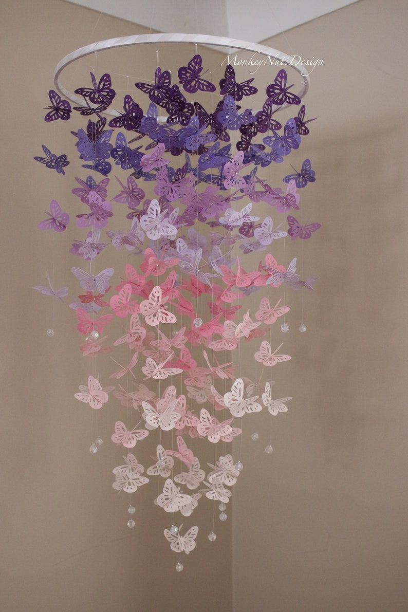 K&ouml;nnte beinhalten: Ein Papier-Schmetterlings-Mobile mit einem Farbverlauf von lila, rosa und wei&szlig;en Schmetterlingen, die an einem wei&szlig;en Metallring h&auml;ngen. Die Schmetterlinge sind in Reihen angeordnet, wobei die dunkelsten lila Schmetterlinge oben und die hellsten wei&szlig;en Schmetterlinge unten sind. Das Mobile ist mit kleinen, klaren Perlen verziert.