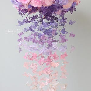 K&ouml;nnte beinhalten: Ein Mobile aus Papier-Schmetterlingen in einem Farbverlauf von Lila &uuml;ber Rosa zu Wei&szlig;. Das Mobile ist mit einem Blumenstrau&szlig; an der Oberseite verziert und hat ein zartes, verspieltes Design.
