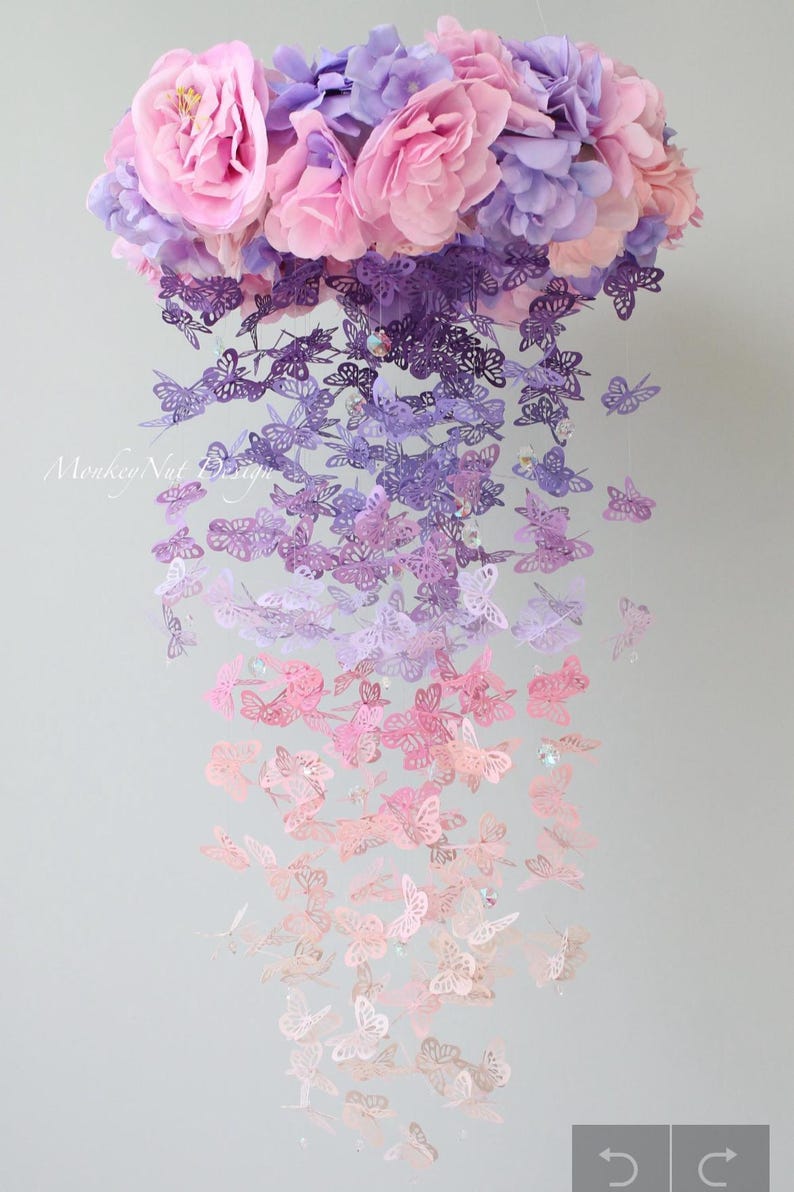 K&ouml;nnte beinhalten: Ein Mobile aus Papier-Schmetterlingen in einem Farbverlauf von Lila &uuml;ber Rosa zu Wei&szlig;. Das Mobile ist mit einem Blumenstrau&szlig; an der Oberseite verziert und hat ein zartes, verspieltes Design.