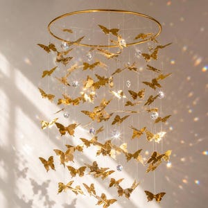 May include: A hanging mobile featuring a circular gold-toned frame with numerous metallic gold butterfly cutouts suspended by thin clear strings. Interspersed among the butterflies are small, clear, faceted crystal beads that catch the light.