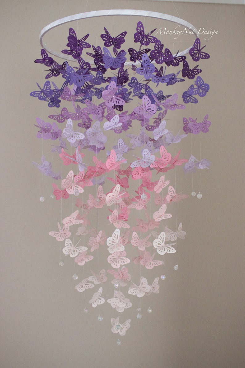K&ouml;nnte beinhalten: Ein Mobile aus Papier in Form von Schmetterlingen in Lila, Lavendel und Rosa mit Kristallperlen, die an einem wei&szlig;en Metallring h&auml;ngen.  "Monkey Nut Design" steht in der oberen rechten Ecke des Bildes.