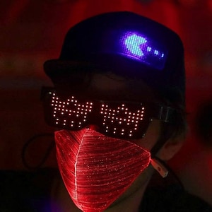 Peut inclure: Une personne portant une casquette noire avec une lumière LED bleue affichant un personnage Pac-Man. Elle porte également un masque lumineux LED rouge avec un motif pixelisé.