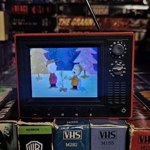 May include: A small, red and black retro-style television displaying a cartoon scene. The TV sits atop a stack of VHS tapes, with titles like "Creepshow" and "The Last Horror" visible. The background is filled with more VHS tapes.