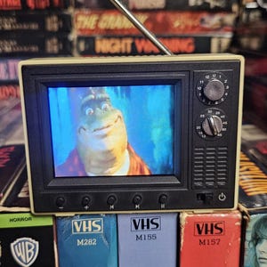 Puede incluir: Un televisor retro en miniatura con una caricatura en la pantalla. El televisor beige tiene controles negros y una antena plateada. Está colocado sobre una pila de cintas VHS, incluyendo "Creepshow" y "Maniac".