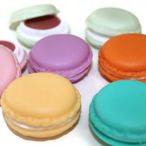 May include: A collection of round, macaron-shaped containers in various colors including pink, purple, orange, teal, yellow, and light green. Some containers are closed, while others are open to reveal a dark interior, arranged on a white surface.
