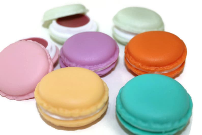 May include: A collection of round, macaron-shaped containers in various colors including pink, purple, orange, teal, yellow, and light green. Some containers are closed, while others are open to reveal a dark interior, arranged on a white surface.