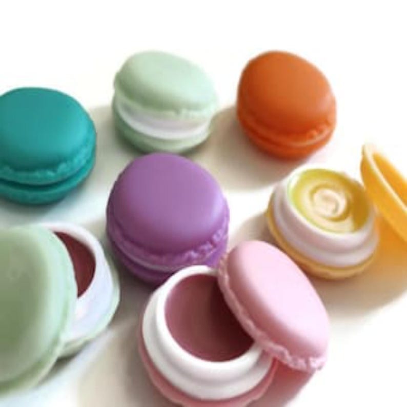 May include: A collection of round, macaron-shaped lip balm containers in various pastel and vibrant colors including teal, light green, orange, purple, pink, and yellow. Some containers are closed, while others are open, revealing colored balm.