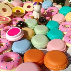 May include: A collection of small, round, colorful plastic containers shaped like donuts and macarons, featuring pastel and bright shades of pink, blue, green, orange, and yellow. A small white unicorn figurine with a purple mane sits among the items.