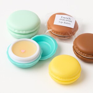 May include: Four round lip balm containers shaped like French macarons in mint green, brown, and yellow. One brown macaron is in clear plastic packaging labeled French macaron Lip Balm. One mint green container is open, revealing yellow balm with a tiny pink heart.