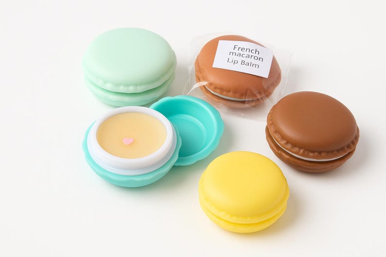 May include: Four round lip balm containers shaped like French macarons in mint green, brown, and yellow. One brown macaron is in clear plastic packaging labeled French macaron Lip Balm. One mint green container is open, revealing yellow balm with a tiny pink heart.