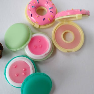 May include: A collection of small, round cosmetic containers shaped like sweets. Included are a pink donut-shaped compact, a dark brown cupcake-shaped container, and two macaron-shaped containers in light green and teal, each holding pink lip balm.