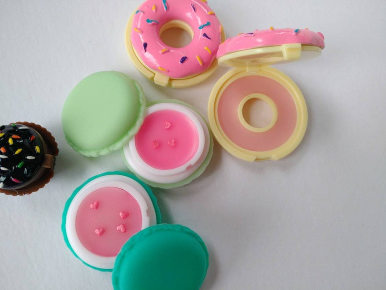 May include: A collection of small, round cosmetic containers shaped like sweets. Included are a pink donut-shaped compact, a dark brown cupcake-shaped container, and two macaron-shaped containers in light green and teal, each holding pink lip balm.