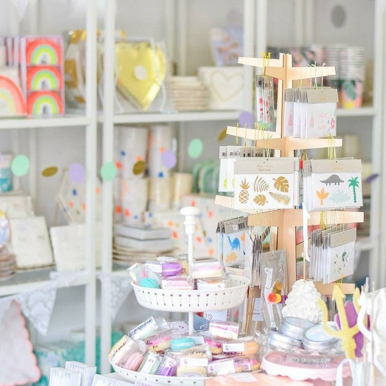 May include: A light-colored wooden display stand holds various hanging packages of temporary tattoos featuring gold foil designs of pineapples, leaves, and dinosaurs, alongside a white tiered tray filled with assorted colorful macarons.