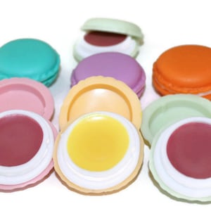 May include: A collection of round, macaron-shaped lip balm containers in various colors including teal, pink, purple, orange, yellow, and light green. Some containers are closed, while others are open, revealing smooth, solid-colored lip balm.