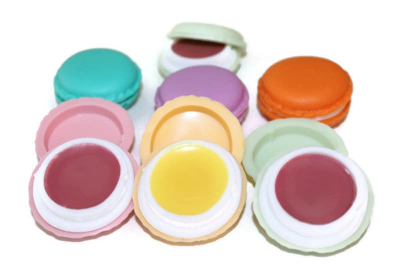 May include: A collection of round, macaron-shaped lip balm containers in various colors including teal, pink, purple, orange, yellow, and light green. Some containers are closed, while others are open, revealing smooth, solid-colored lip balm.