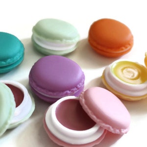 May include: A collection of round, macaron-shaped lip balm containers in various colors including teal, light green, orange, purple, pink, and yellow. Some containers are closed, while others are open, revealing tinted lip balm inside.