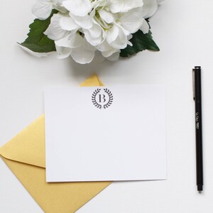 Könnte beinhalten: Ein weißer Umschlag mit einem goldenen Umschlag im Inneren. Der weiße Umschlag hat einen einfachen Kranz mit dem Buchstaben "B" in der Mitte. Ein schwarzer Stift liegt auf der weißen Oberfläche.