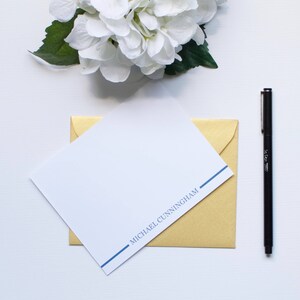 Könnte beinhalten: Eine weiße Karte mit einem blauen Strich und dem Text "MICHAEL CUNNINGHAM" darauf gedruckt. Die Karte liegt auf einem goldenen Umschlag.