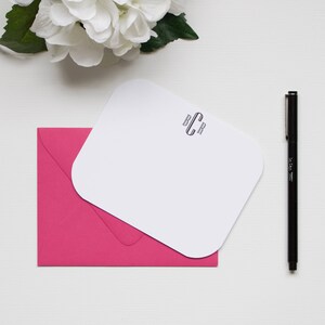 May include: A stationery set featuring a white, rounded-corner notepad with a monogram, resting on a bright pink envelope. A black pen and white flowers are also present, all on a white surface.