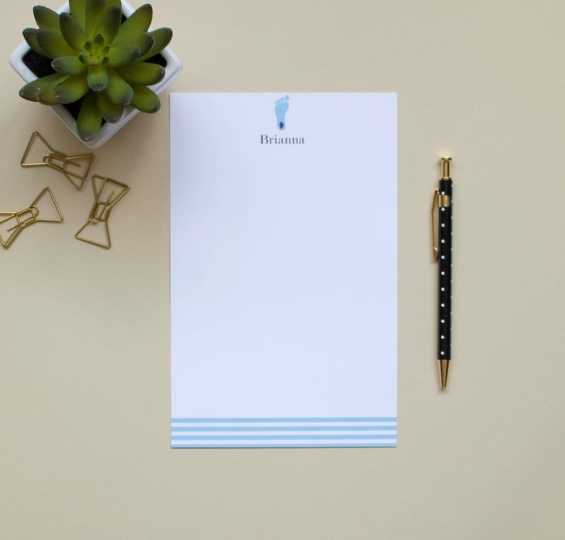 Puede incluir: Una hoja de papel de carta personalizado blanco con rayas horizontales azules en la parte inferior. El nombre "Brianna" est&aacute; impreso en negro en la parte superior de la hoja, con un icono de huella azul sobre el nombre. Un bol&iacute;grafo negro con lunares blancos est&aacute; sobre una superficie beige junto al papel de carta.