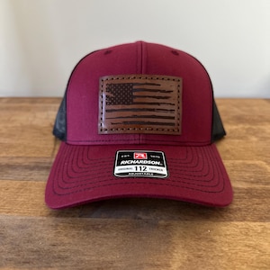 Puede incluir: Una gorra de camionero de color burdeos y negro con un parche de cuero marrón que presenta una bandera estadounidense. La gorra tiene una correa ajustable y una etiqueta que dice "Richardson. Original 112 Trucker Adjustable."