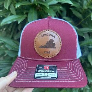 May include: A maroon and white trucker hat with a brown leather patch featuring the state of Virginia and the text "VIRGINIA 1788". The hat is adjustable and has the Richardson logo and the text "EST. 1970 ORIGINAL 112 TRUCKER ADJUSTABLE".