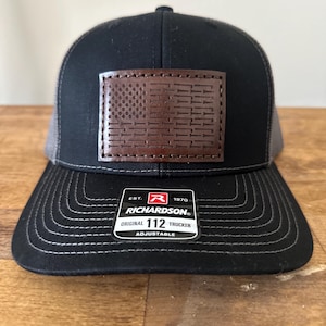May include: Black and gray trucker hat with a brown leather patch featuring an American flag design. The patch has a stitched border and the text "EST. 1970 RICHARDSON ORIGINAL 112 TRUCKER ADJUSTABLE" is printed on a white label attached to the hat.