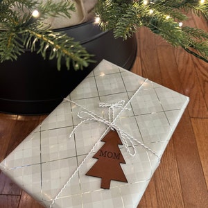 Op de afbeelding: Een witte cadeaudoos met een zilveren ruitpatroon is ingepakt met wit touw en een bruine leren kerstboomhanger met de tekst "MOM".