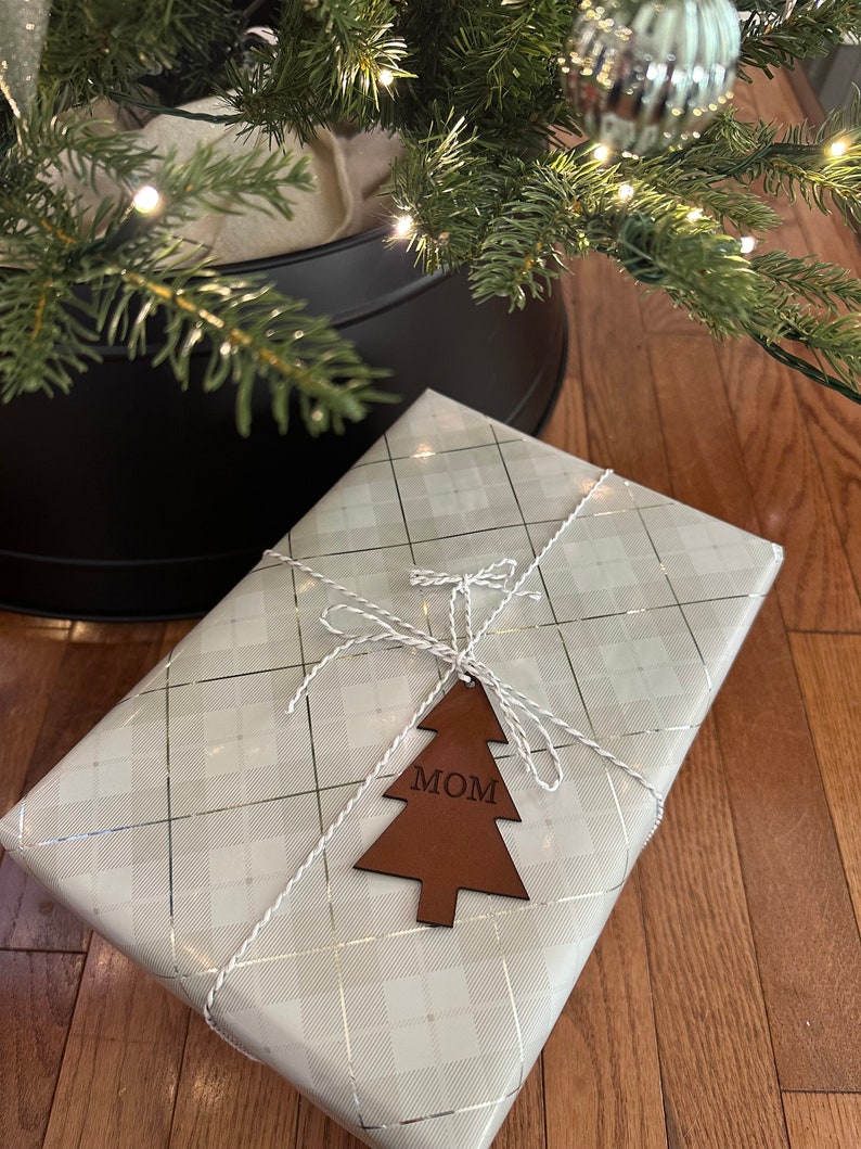 Op de afbeelding: Een witte cadeaudoos met een zilveren ruitpatroon is ingepakt met wit touw en een bruine leren kerstboomhanger met de tekst "MOM".