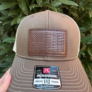 Puede incluir: Una gorra de camionero marrón y blanca con un parche de cuero marrón que presenta un diseño de bandera estadounidense. El parche tiene un borde cosido y la gorra tiene una parte posterior de malla blanca. La gorra tiene una etiqueta que dice "EST. 1970 RICHARDSON ORIGINAL 112 TRUCKER ADJUSTABLE".