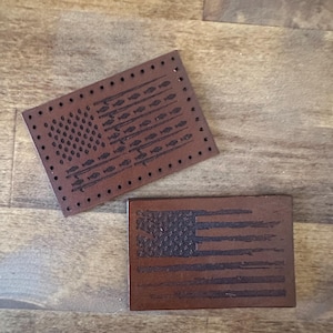May include: Two brown leather patches with an American flag design. One patch features a traditional flag design with stars and stripes, while the other patch features a more stylized flag design with fishing rods and fish.