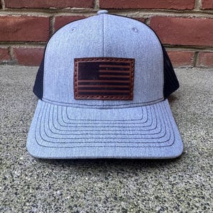 Könnte beinhalten: Eine graue und schwarze Trucker-Kappe mit einem braunen Leder-Patch der amerikanischen Flagge.