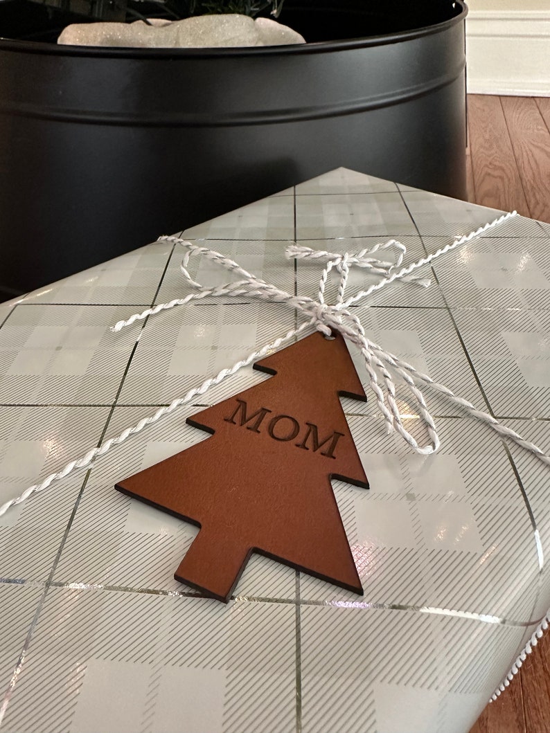 Op de afbeelding: Een bruine houten kerstboomhanger met het woord "MOM" erop gegraveerd. De hanger is vastgebonden met wit touw en ligt op wit en grijs geruit cadeaupapier.
