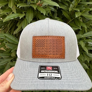 Puede incluir: Gorra de camionero gris y blanca con un parche de cuero marrón que presenta un diseño de bandera estadounidense estilizado. El parche tiene el texto "Richardson" y "112". La gorra es ajustable.