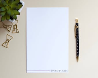 Op de afbeelding: Een wit vel papier met een zwarte lijn aan de onderkant en de tekst "RODNEY D. ROLFE" in zwart gedrukt. Een zwarte pen met witte stippen bevindt zich aan de rechterkant van het papier. Aan de linkerkant van het papier bevinden zich vier gouden paperclips in de vorm van een strik.