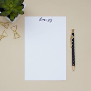 Puede incluir: Una hoja de papel blanca con las palabras "choose joy" escritas en tinta morada. Un bolígrafo negro con lunares blancos está sobre una superficie beige junto al papel.