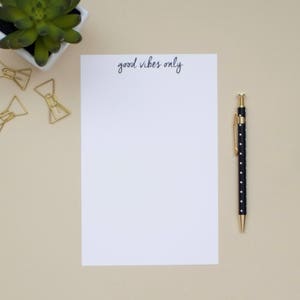 May include: A white notepad with the text "good vibes only" written in black ink. A black pen with white polka dots is on the right side of the notepad. There are gold paper clips on the left side of the notepad.
