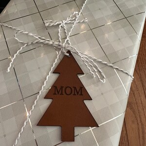 Op de afbeelding: Een bruine leren kerstboomhanger met het woord "MOM" erop gegraveerd. De hanger is vastgebonden met wit touw aan wit en zilver cadeaupapier.