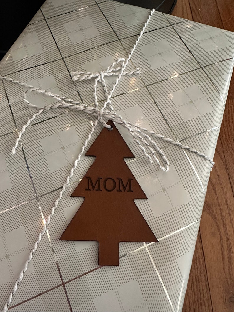 Op de afbeelding: Een bruine leren kerstboomhanger met het woord "MOM" erop gegraveerd. De hanger is vastgebonden met wit touw aan wit en zilver cadeaupapier.