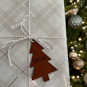 Op de afbeelding: Een witte cadeaudoos met een zilver en wit geruit patroon is ingepakt met witte touw en een bruin houten kerstboom ornament met de tekst "MOM".