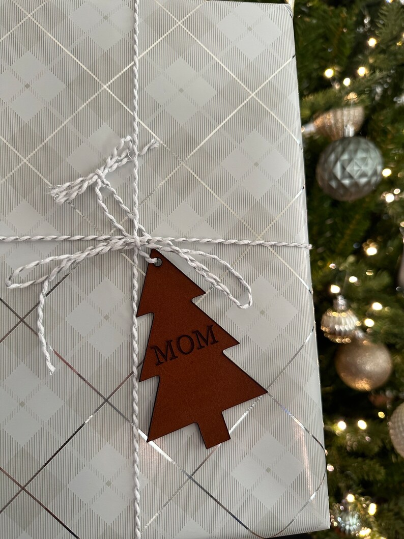 Op de afbeelding: Een witte cadeaudoos met een zilver en wit geruit patroon is ingepakt met witte touw en een bruin houten kerstboom ornament met de tekst "MOM".