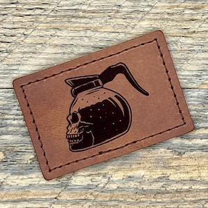 May include: A brown leather patch with a stitched border. The patch features a black graphic of a coffee pot in the shape of a skull. The patch is rectangular and has a rustic, handmade appearance.