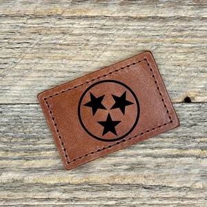 May include: A brown leather patch with a stitched border featuring the state of Tennessee's flag, a white circle with three black stars.